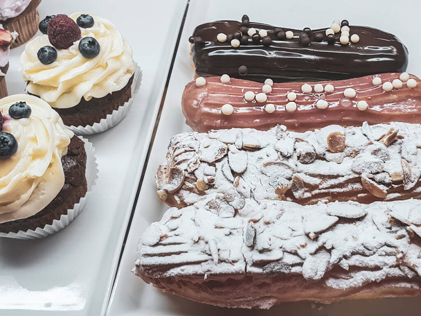 застекленные эклеры и кексы со сливками и ягодами за стеклянным футляром, сладкий десерт
 - Фото, изображение