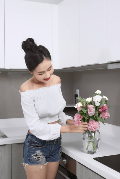 Nuori kaunis aasialainen nainen järjestää kukkia maljakko keittiössä
. - Valokuva, kuva