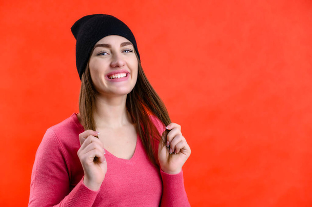 Portrait of a pretty young woman with a smile in a hat in a pink sweater on a red background with joyful emotions - 写真・画像