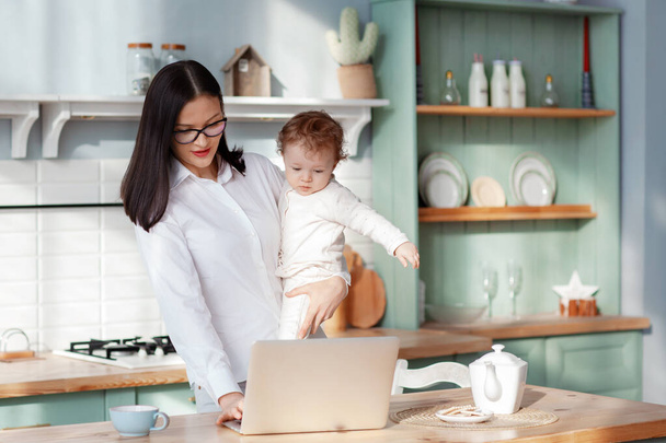 Jovem mãe trabalha em casa com um laptop com um bebê nos braços, trabalho remoto em licença maternidade
 - Foto, Imagem