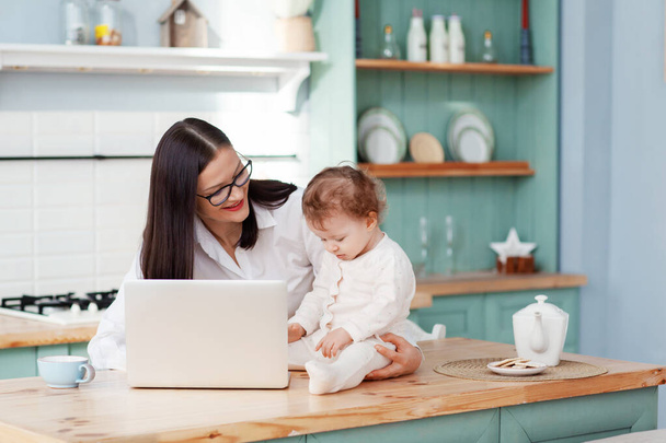 Jovem mãe trabalha em casa com um laptop com um bebê nos braços, trabalho remoto em licença maternidade
 - Foto, Imagem