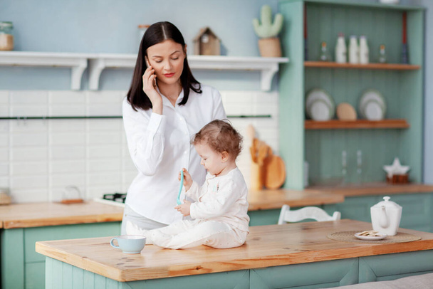 Młoda matka rozmawiająca przez telefon w domu w kuchni z dzieckiem w ramionach, zdalna praca na urlopie macierzyńskim - Zdjęcie, obraz