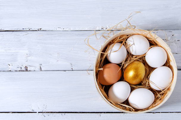 Ιστορικό Πάσχα. Σε ένα κόσκινο στο σανό υπάρχουν λευκά αυγά κότας, ένα χρυσό αυγό, ένα καφέ. Ένα κόσκινο στέκεται σε ένα ξύλινο λευκό φόντο. Επίπεδη διάταξη, οριζόντιο φόντο - Φωτογραφία, εικόνα