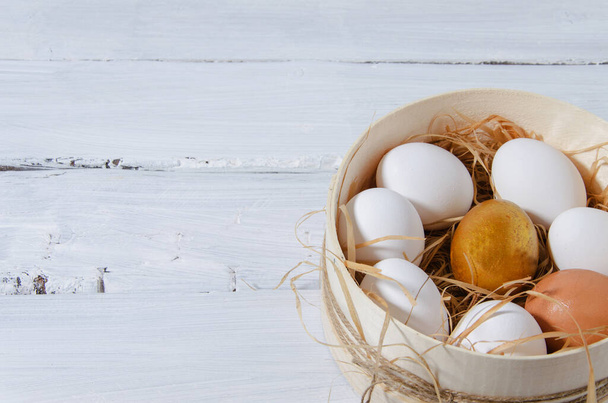 Fondo de Pascua. En un tamiz en el heno hay huevos de gallina blanca, un huevo de oro, uno marrón. Un tamiz está sobre un fondo blanco de madera. fondo horizontal
 - Foto, imagen