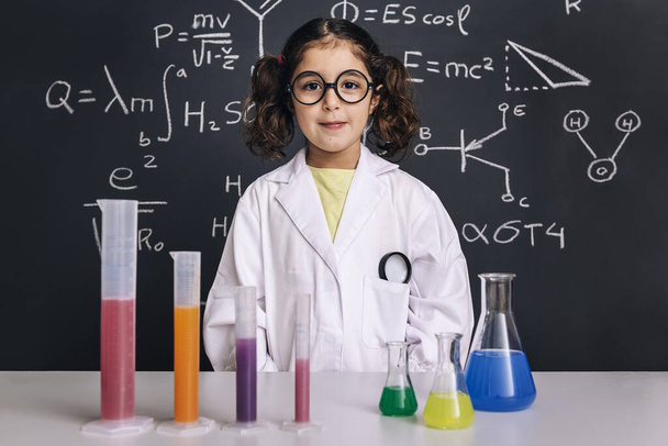 little scientist girl with glasses in lab coat with chemical flasks, on school blackboard background with hand drawn science formulas, back to school and successful female career concept - Φωτογραφία, εικόνα