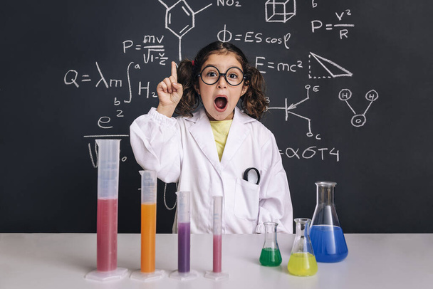 surprised scientist child with glasses in lab coat with chemical flasks having an idea, school blackboard background with science formulas, back to school and successful female career concept - Foto, afbeelding