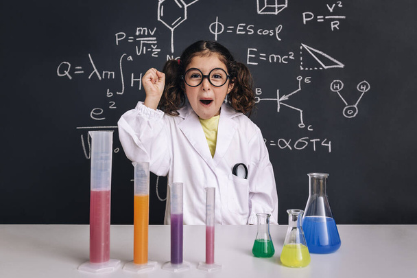 happy scientist child with glasses in lab coat with chemical flasks celebrating an idea, school blackboard background with science formulas, back to school and successful female career concept - Photo, image