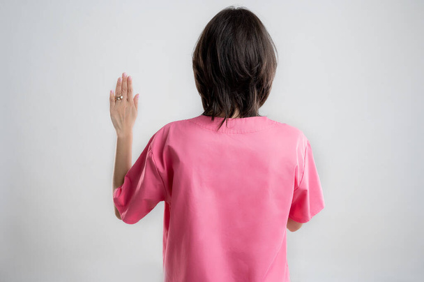 Jonge vrouw medische beroepsverpleegkundige of arts gekleed met roze ziekenhuis kleding, met bruin haar, tonen eed van achteren poseren op een witte geïsoleerde back-round. - Foto, afbeelding