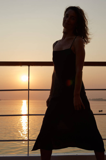 Sinnlich authentische Aufnahme einer attraktiven jungen kaukasischen Frau im langen schwarzen Kleid, die an Bord eines Kreuzfahrtschiffes posiert, sich glücklich und zuversichtlich fühlt und den malerischen Sonnenuntergang mit herrlichem Blick auf den Fluss genießt. - Foto, Bild