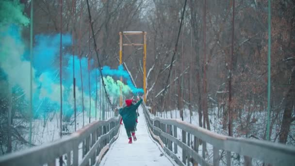 Two young women running on the snowy bridge holding smoke bombs - Πλάνα, βίντεο