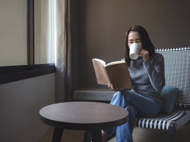 Azja piękna kobieta w ubraniu casual czytania książki z napojem na krześle w pokoju z miejsca do kopiowania. Miękkie skupienie. - Zdjęcie, obraz
