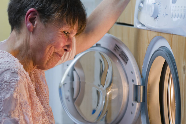 洗濯機の開口部に満足そうに見ている高齢女性の側面の頭と肩のビュー. - 写真・画像