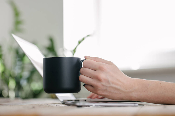 Schwarze Metalltasse für Tee und Kaffee in Frauenhänden im Büro. Sie benutzt Laptop und trinkt Kaffee aus schwarzem Metallbecher - Foto, Bild