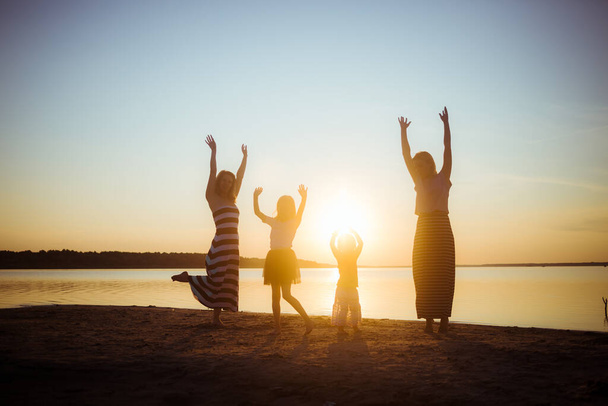Силуэты детей и их матерей, прыгающих и веселящихся на пляже в лучах заката. Хорошее настроение и развлечение среди молодого и старшего поколения. Красивый пейзаж. - Фото, изображение