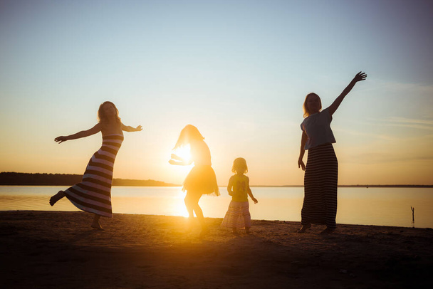 Silhouette di bambini e le loro madri che saltano e si divertono sulla spiaggia alla luce del tramonto. Buon umore e passatempo tra le generazioni più giovani e più anziane. Bellissimo paesaggio. - Foto, immagini