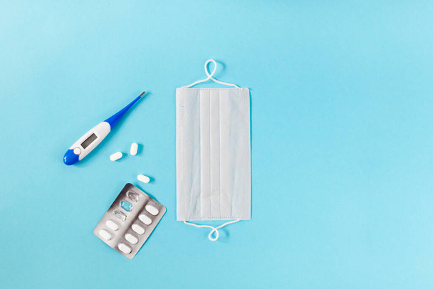 Fehér orvosi védőmaszk, pirulákkal és hőmérővel kék háttérrel. Coronovírus megelőzés és egészségügyi koncepció. horizontális nézet copyspace - Fotó, kép