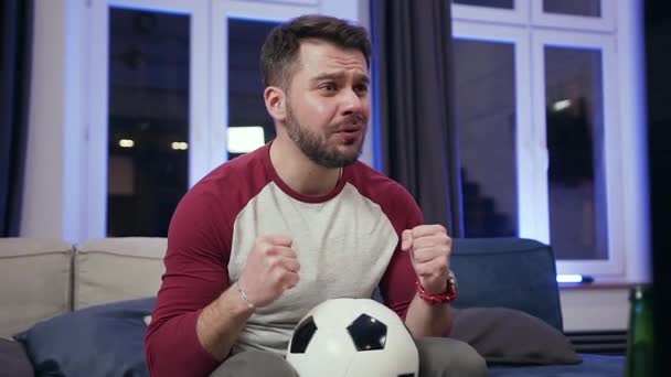 Attractive excited bearded young guy holding ball in his hands and shouting "goal "during watching football match - Filmagem, Vídeo