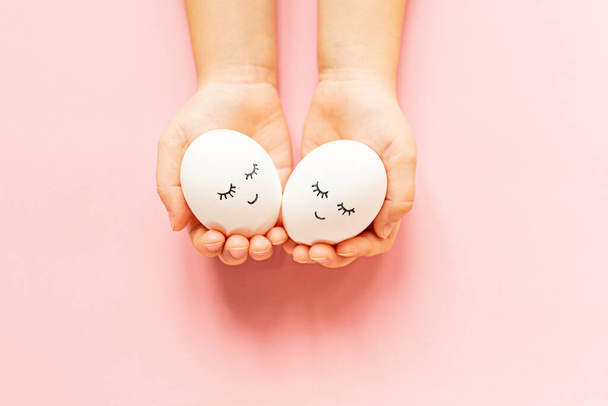 Hands holding easter eggs with cute face and sleepy eyes on pastel pink background. Happy Easter concept. Flat lay, top view, mockup, template, overhead, copy space. - Φωτογραφία, εικόνα