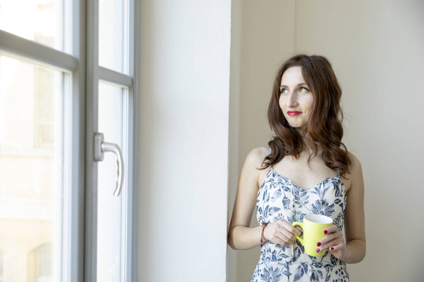 Brunette girl with long hair looks out the window with a yellow cup of tea. She looks out. - Foto, Imagen