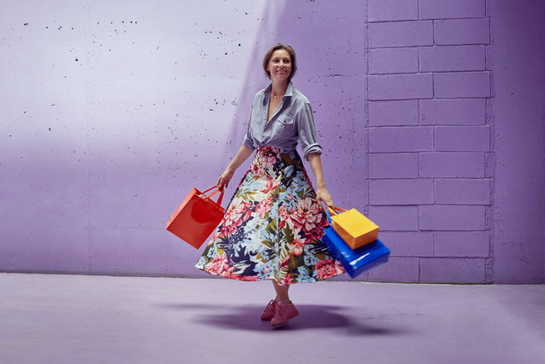 Νεαρό όμορφο κορίτσι με πακέτα ψώνια στα χέρια. Ένα κορίτσι με μια μακριά φωτεινή φούστα και ανοιχτό ροζ μπλουζάκι περπατά από το κατάστημα. - Φωτογραφία, εικόνα