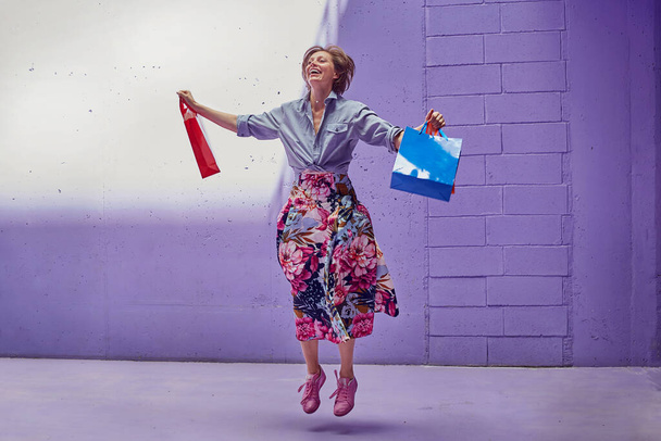 Young beautiful girl jumping of joy with shopping in her hands. A girl in a long bright skirt and pale pink top is walking from the store. - Foto, imagen