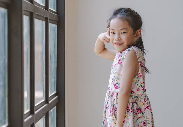 Cute act pose portrait of Asian little girl, standing pose next to big window, act two fingers on face, smiling face and looking at camera, wearing flower pattern dress. Confident and fun emotion. - Foto, afbeelding