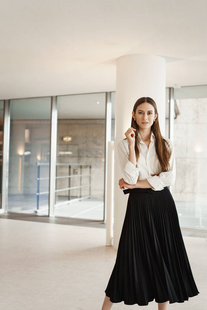 Vertical portrait of independent strong woman in black formal skirt and white blouse, working in office, standing confident in white corridor, expressing self-assurance and success - 写真・画像