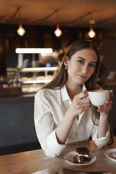Vertical portrait of romantic feminine young woman drinking coffee alone in cozy cafe, leaning elbows wooden table, holding cup and smiling at camera, eating cake during lunch - Valokuva, kuva
