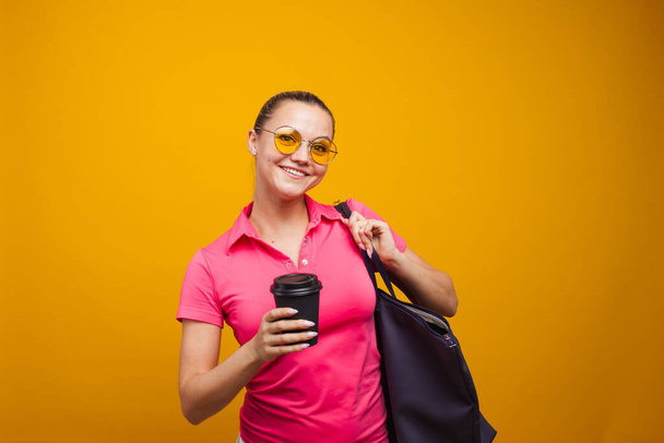 Cute blonde in sunglasses and t-shirt with bag on shoulder, drinking her hot drink, portrait on yellow background - Fotografie, Obrázek