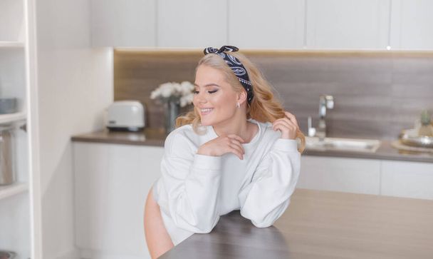 Seksikäs hymyilevä blondi otsanauhassa poseeraa lähellä keittiön pöytää valkoisessa puserossa ja pikkuhousuissa
 - Valokuva, kuva