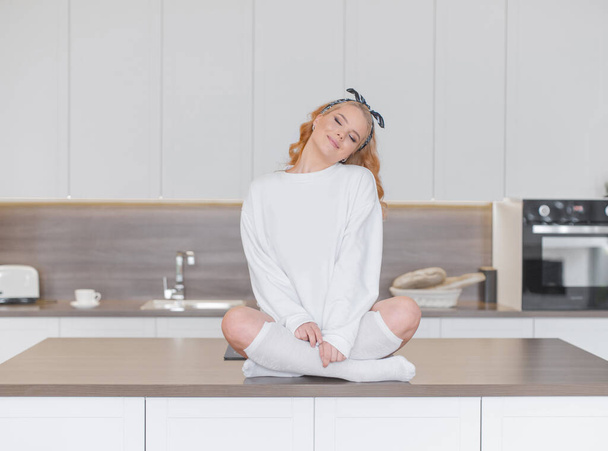 Sexy blond in wit sweatshirt en knie sokken zitten in Turkse pose op de keukentafel - Foto, afbeelding