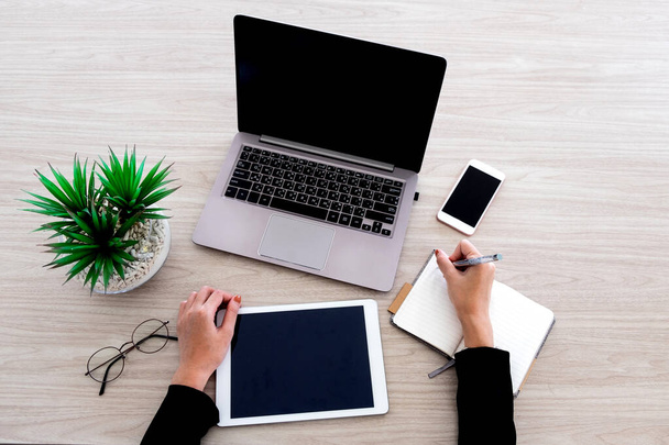 Женщина работает с различными офисными устройствами, такими как ноутбук, планшет и смартфон. Она пишет на маленьком блокноте на деревянном столе в офисе
. - Фото, изображение