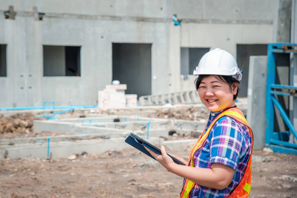 Ázsiai nő építőmérnök vagy építész sisak és biztonsági mellény dolgozik, és a kezében egy érintésmentes táblagép látni tervrajzok vagy terv egy épület vagy építési területen - Fotó, kép