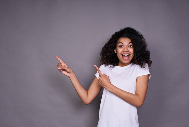 A cute beautiful cheerful young dark-skinned girl, a student, in a white basic T-shirt, with black curly hair, shows his fingers to the left, expresses joy and surprise. - Photo, image
