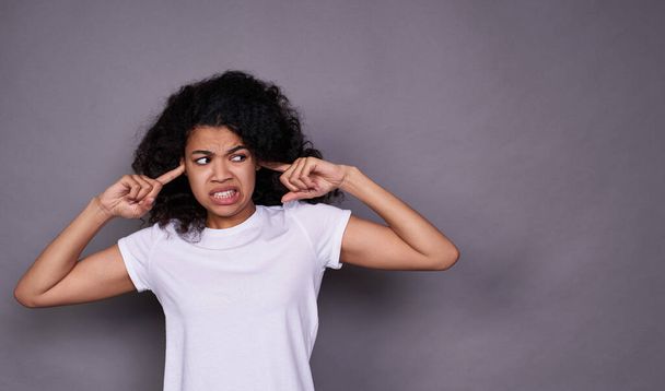 Frustrated, angry, young dark-skinned girl with black curly hair, in a white basic T-shirt, disgusted, covers her ears, does not want to hear, looks to the right. - Photo, Image