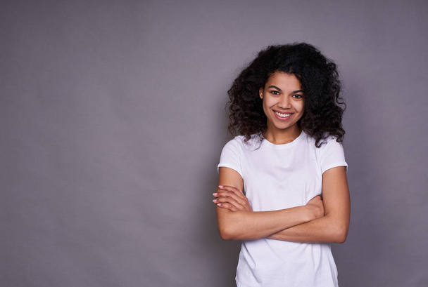 A cheerful African American with a pleasant smile, curly dark hair, healthy skin, wears a white T-shirt, stands on a gray background, being in a good mood, crossed her arms over her chest. - Foto, Imagen