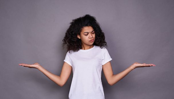 A young cute cute African woman, about 20 years old, in a white basic T-shirt, standing on a gray background, making a choice, spread her arms to the side, looking to the side. - Foto, imagen