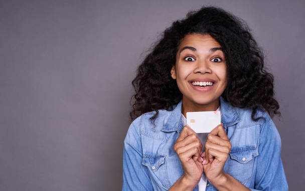 Atractiva joven africana en una camiseta blanca y camisa de mezclilla con su tarjeta de crédito estirándola a la cámara, la cabeza y los hombros, sonriendo felizmente y mirando recto
. - Foto, Imagen
