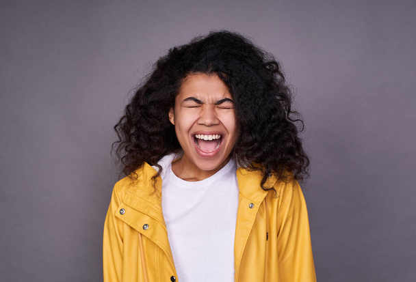 Young pretty funny, funny dark-skinned girl with a lush curly hair, in a white T-shirt and yellow raincoat, closed her eyes and opened her mouth in a cry, photo on a gray background. - Foto, afbeelding