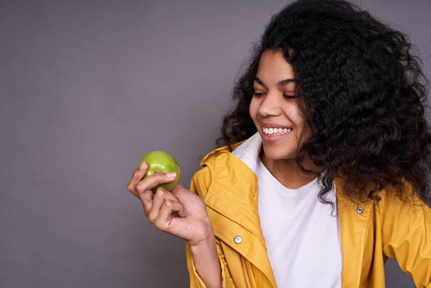 Close-up, joyful funny dark-skinned young lady, with curly hair, in a white T-shirt and a raincoat token, holds a green apple in his hand and laughs, looks at the apple in his right hand. - Photo, Image