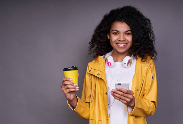 A young pretty gentle Afrikaan girl with a lush curly hairdo, in a white T-shirt and yellow jacket, holds a coffee cup in her hand and looks straight at smiling. - Photo, image