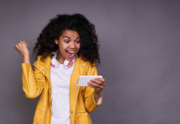 Cute cheerful happy young African girl, in a white T-shirt, with magnificent curly hair, holds a smartphone in her hand, learns good news, good luck, victory. Photo isolated on a gray background. - Φωτογραφία, εικόνα