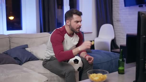 Handsome concentrated bearded young man eating chipps during reviewing the football match on tv at home - Filmagem, Vídeo