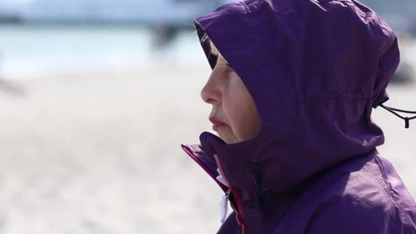 Dorosła kobieta w fioletowej kurtce i kapturze zjada kanapkę siedzącą na plaży w słoneczny dzień, wiosną - Materiał filmowy, wideo