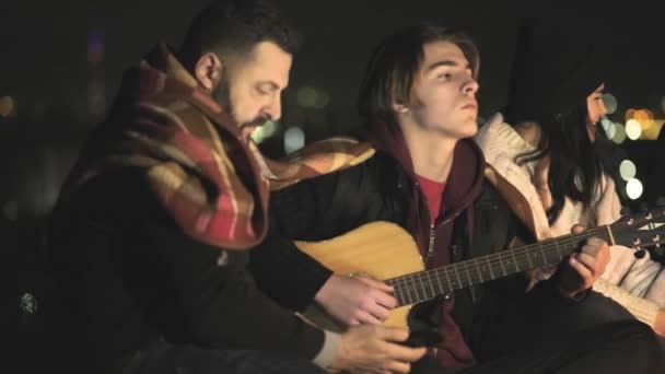 Grupa ludzi bawiących się siedząc w pobliżu ogniska na zewnątrz w nocy grając na gitarze, śpiewając piosenki i rozmawiając szczęśliwie razem. - Materiał filmowy, wideo