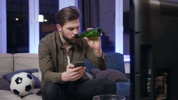 Front view of concentrated handsome young bearded man which drinking beer while browsing his phone in front of tv during sport game - 映像、動画