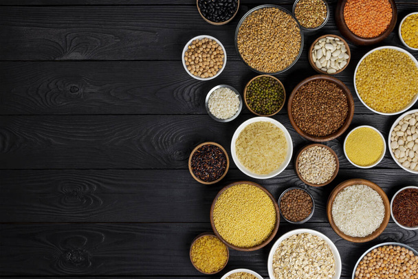 Různé obiloviny, zrna, semena, krupice, luštěniny a fazole v miskách na černém dřevěném pozadí s kopírovacím prostorem - Fotografie, Obrázek