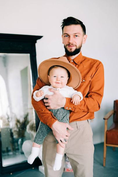 a young stylish father in an orange shirt plays with his young son in a brown hat in his arms - Photo, image