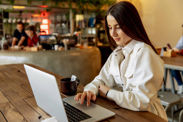 Charming female freelancer in trendy clothing working on laptop during morning breakfast at cozy cafe. Pretty young girls with brown hair smiling and posing on camera. - Φωτογραφία, εικόνα