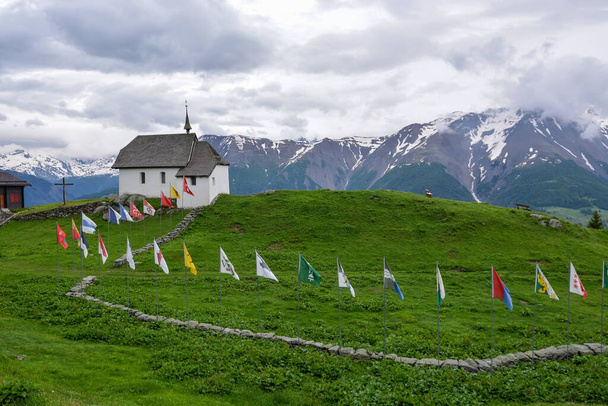 Церковь с большим количеством флагов в швейцарской деревне Бетмеральп в пасмурный день
 - Фото, изображение